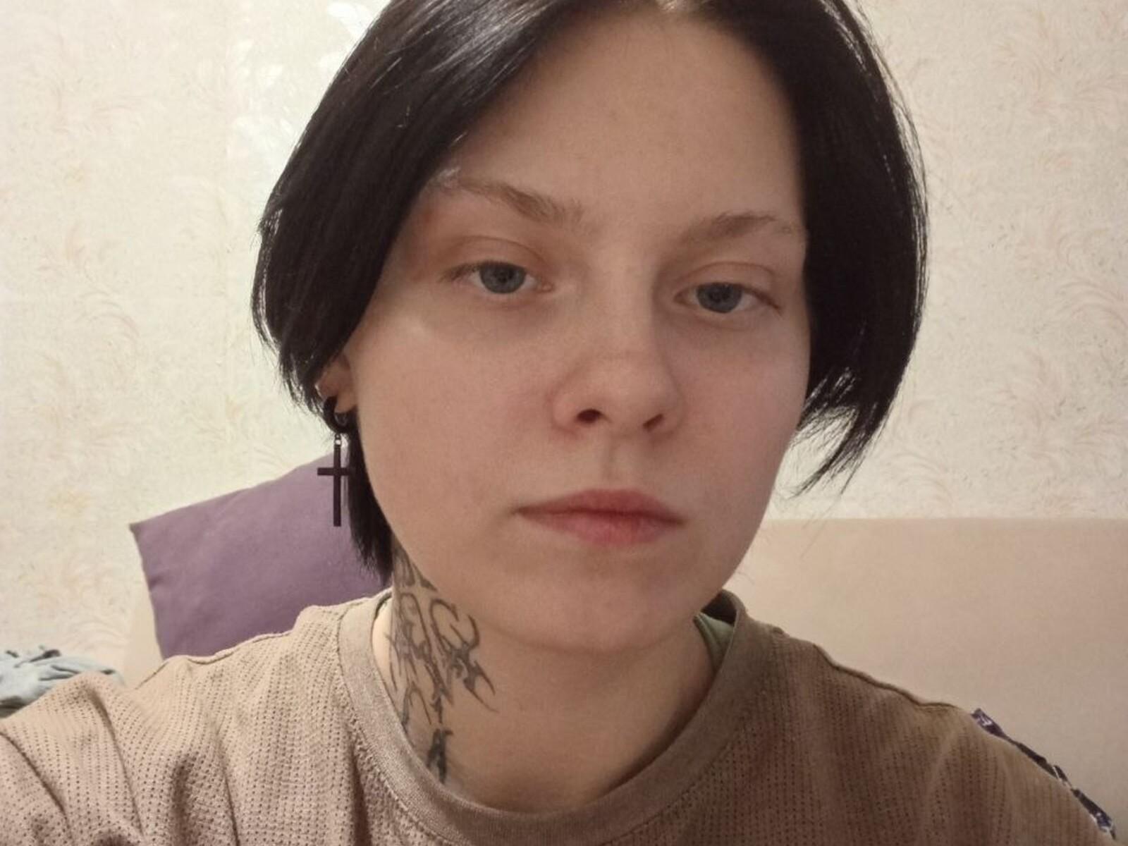 LuellaIshi met kort zwart haar, bleke huid, een kleine tatoeage op de nek, een kruisje als oorbel, een neutrale uitdrukking, een beige trui, geen make-up en een lichtjes fronsende gezichtsuitdrukking..