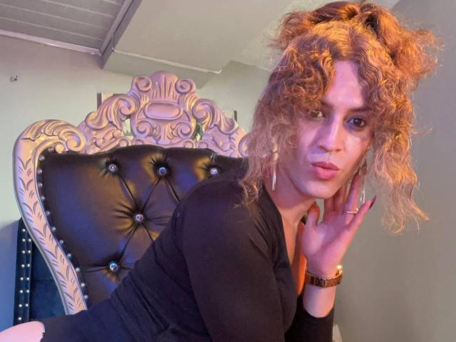 RossSofiia with curly blonde hair, black top, and gold earrings, pouting with a hand near face, sitting on a tufted, black, ornate chair, wearing a wristwatch.