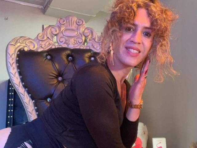 RossSofiia with curly blonde hair, black long-sleeve shirt, and gold earrings, leaning on a tufted black leather chair, smiling, showing teeth, hand on cheek.