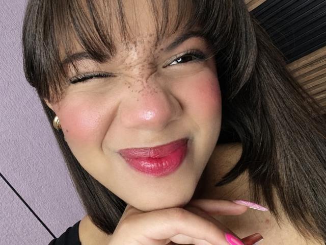 ZaraBlaze has brown hair, freckles, and pink lipstick, winking with a smile, showing off her shiny nose and pink nails, touching her chin with a cute, flirty look.
