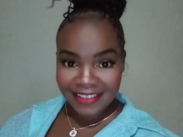 PinkyPiexx has dark skin, curly updo, thick eyebrows, red lipstick, blue jacket, silver necklace with flower pendant, smiling, looking at camera.
