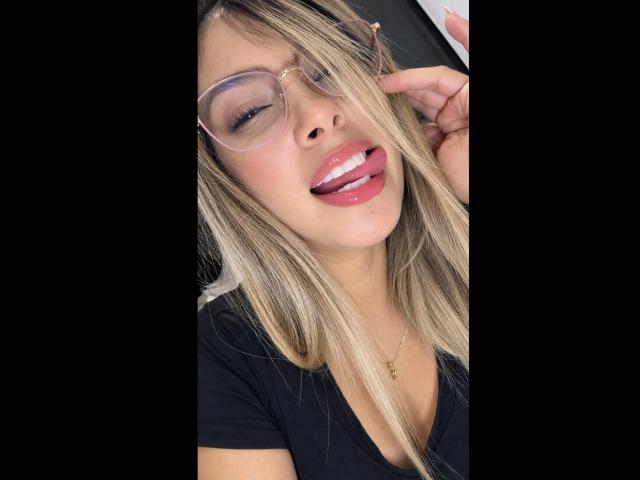 KendraMiler's face, blonde hair, wearing glasses, black top, tongue out, showing white braces, gold necklace, hand touching glasses, black background, slightly smiling, looking cute.