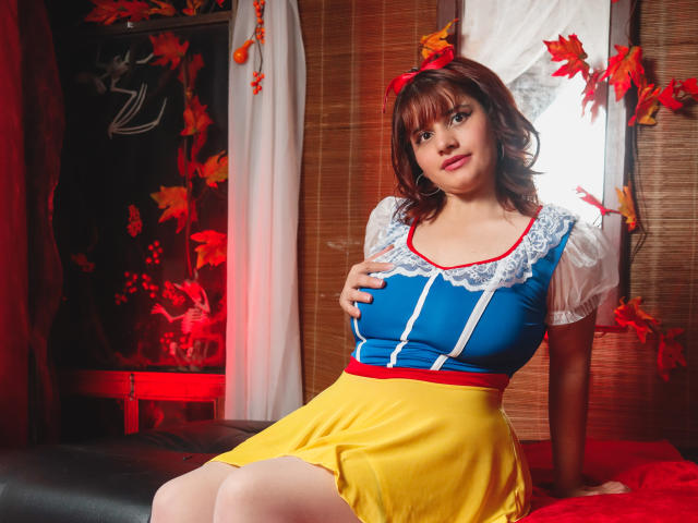 VivianFranco in a Snow White cosplay, wearing a blue top with white lace, yellow skirt, red ribbon, and brown wig, sitting with hand on her tits, red leaves in background.