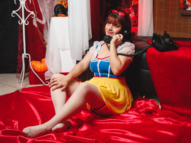 VivianFranco with brown hair, wearing a blue and yellow dress, sitting on red satin, talking on a black phone, barefoot, her tits visible, red ribbon in her hair.