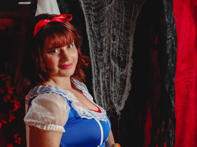 VivianFranco with red hair, blue dress, white lace sleeves, and red bow, smiling at camera, showing her tits, standing in front of black and red fabric.