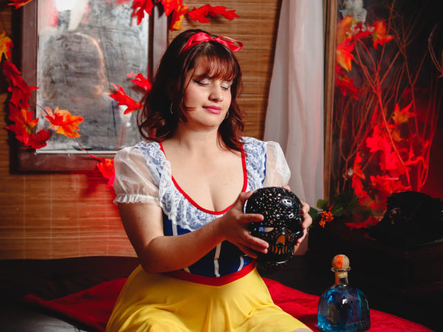 VivianFranco with brown hair, red ribbon, blue and white dress, yellow skirt, holding a black ballerina mask, red and orange leaves, sitting on red cloth, blue bottle.