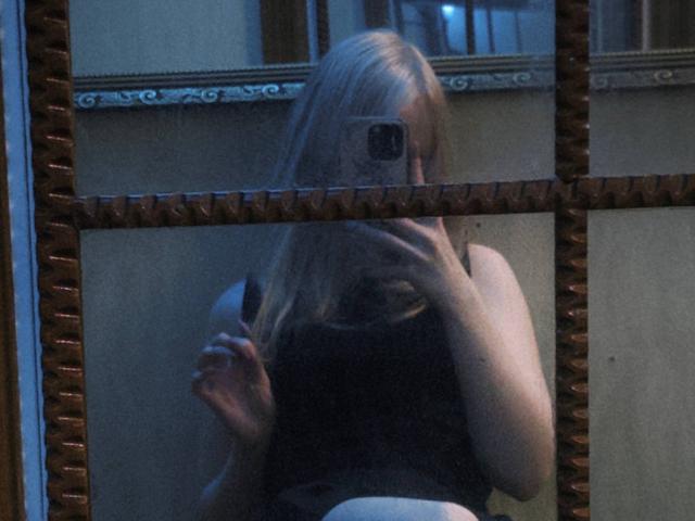 Selfie en el espejo de TiffaniMacandog con cabello rubio, camiseta negra, ropa interior blanca, sosteniendo el teléfono, cubriendo su rostro, reflejada en un espejo ornamentado, mostrando senos pequeños y un ligero trasero..