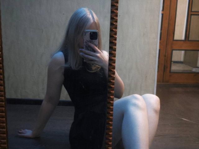 TiffaniMacandog, con cabello plateado, vestido negro sin mangas y medias blancas, sentada en el suelo de madera, tomando una selfie con el teléfono, con las piernas cruzadas y los brazos detrás..