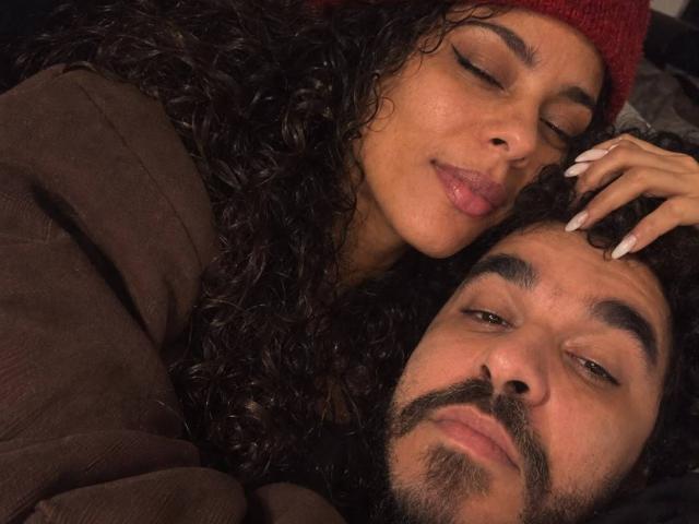 GabyAndVictor with curly brown hair, red beanie, and long nails, resting her head on Victor's curly brown beard, both with closed eyes, lying down, brown jacket.