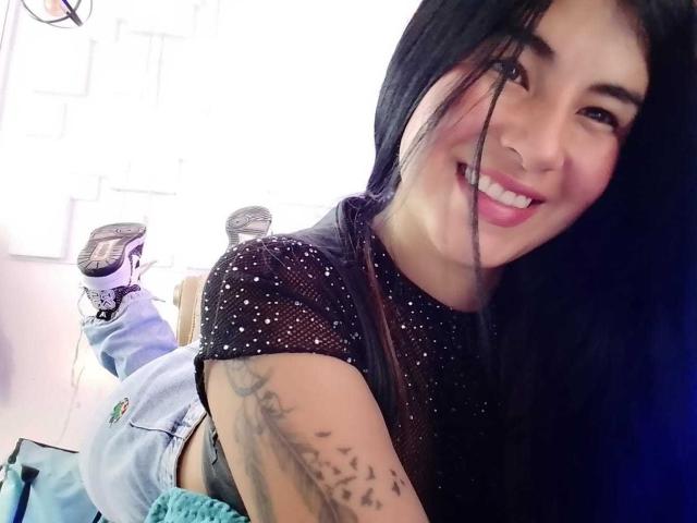 NinaHarpper with long black hair, smiling, wearing a sparkly black top, showing a bird tattoo on her arm, and blue jeans, lying on a couch with legs up.