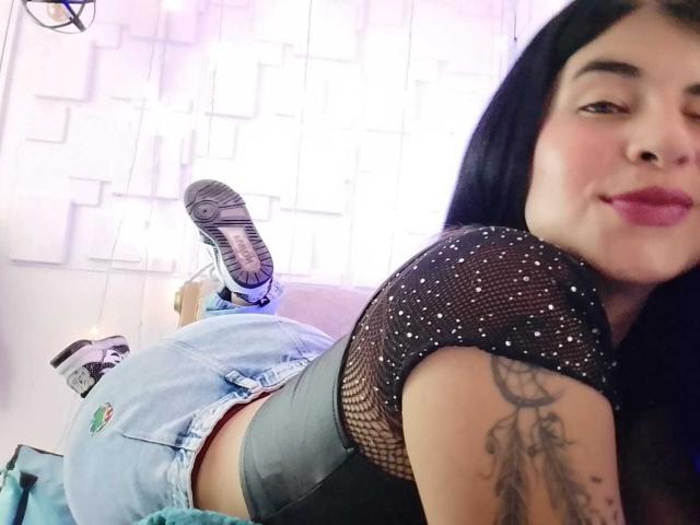 NinaHarpper with long black hair, black mesh top, blue jeans, gray underwear, and white sneakers, lying on her stomach, showing her tattooed arm and smiling at the camera.