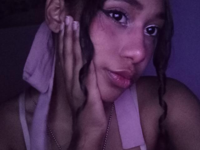 LovelyConnie with braided hair, wearing a white tank top, holding a towel against her face, has a pensive expression, pinkish-purple lighting, and a necklace with a ring.