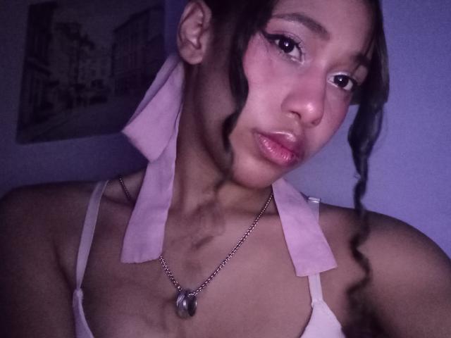 LovelyConnie with dark hair in a ponytail, wearing a pink towel over her shoulders, white bra, and necklace with a heart pendant, looking at the camera with glossy lips.