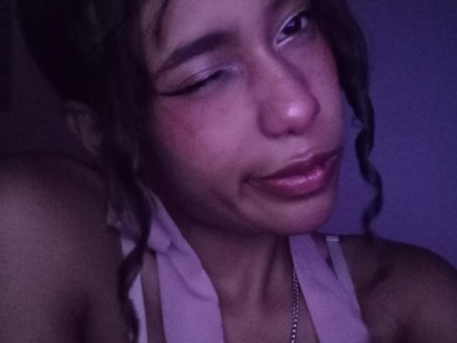 LovelyConnie has one eye closed, smiling with a slightly flushed face, dark curly hair, and is wearing a pink halter top, showing a hint of a necklace.