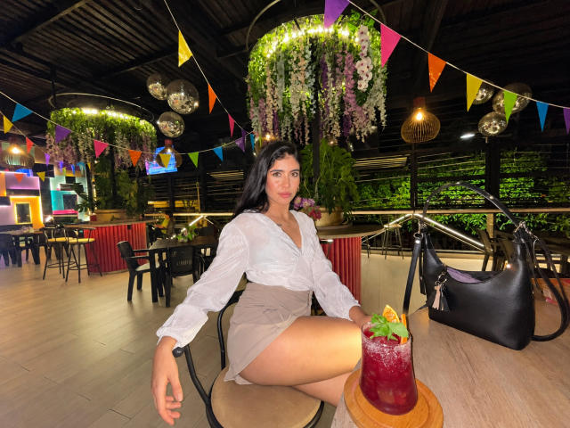 MilusSky com atributos físicos marcantes, vestindo uma blusa branca e uma saia bege, sentada em uma cadeira, segurando uma bebida com frutas, uma bolsa preta no chão, bandeiras coloridas, flores penduradas e luzes no teto..
