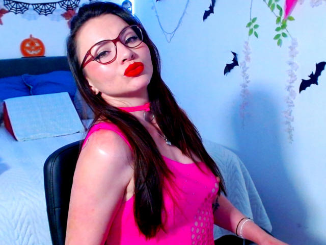 EmelySofia with long brown hair, red lipstick, glasses, pink choker, and pink tank top, sitting in a chair, showing her shiny, slightly exposed tits.