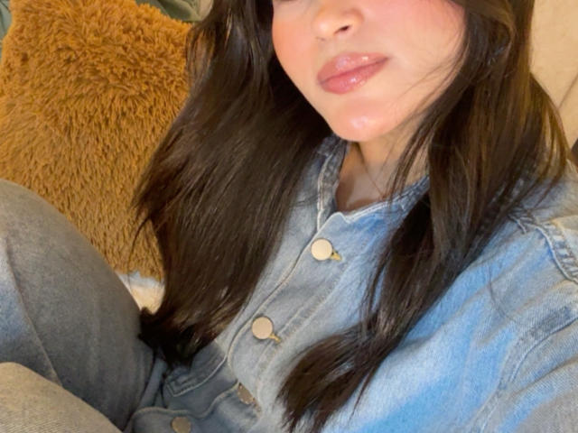 Miia Palmer, con labios brillantes, lleva una camisa vaquera azul y pantalones grises. Tiene el pelo largo y negro, y en el fondo se ve un cojín marrón y esponjoso. Sus pechos son parcialmente visibles..