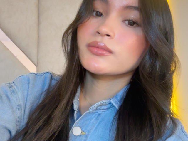 MiiaPalmer tiene labios rosados y brillantes, cabello castaño largo y ondulado, y lleva una camisa vaquera azul con botones visibles. Su piel es clara con un ligero rubor..