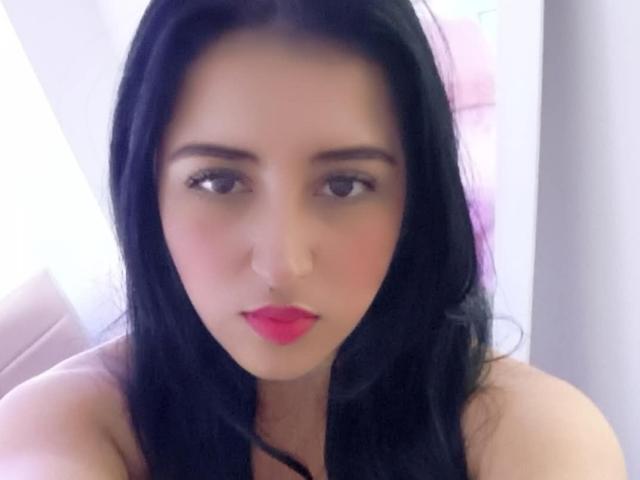 NicollJeans has long black hair, pink lipstick, and a serious expression. Her brown eyes are slightly downcast. She's topless, showing her bare shoulders and soft skin.