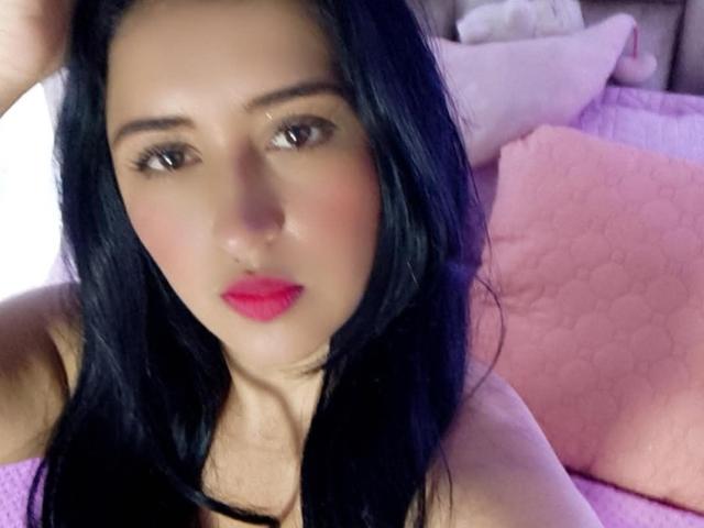 NicollJeans has black hair, pink lipstick, and a cute face with brown eyes, looking at the camera with a slight pout, her hand in her hair. She's topless.