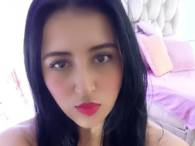 NicollJeans has black hair, brown eyes, red lipstick, and a fair skin tone. She's looking at the camera with a neutral expression. Her tits are not visible.