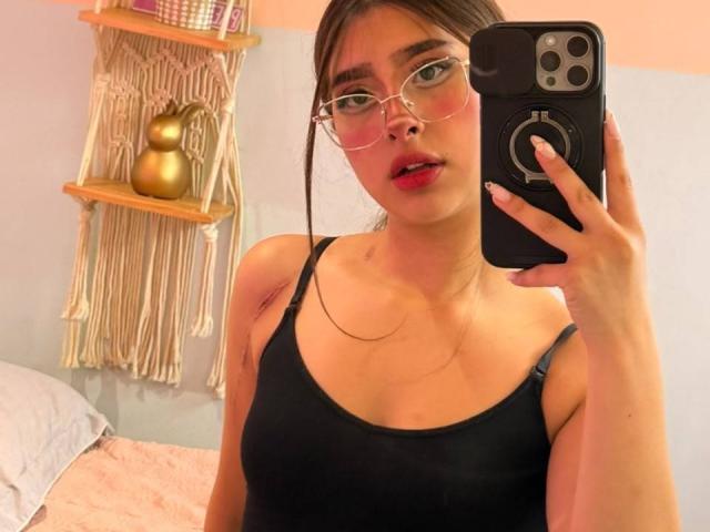 MelanyBlom with glasses, red lipstick, and black tank top, holding a phone in a mirror selfie, showing her tits and face. Her phone has a ring grip.