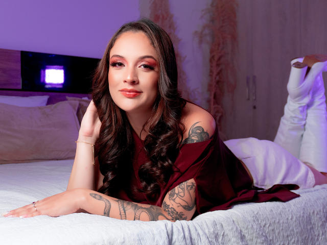 LizethVerda, com cabelos castanhos longos, maquiagem vermelha e tatuagens, deitada em uma cama usando um vestido vermelho, sorrindo com batom vermelho, mão direita na cama, braço esquerdo visível..