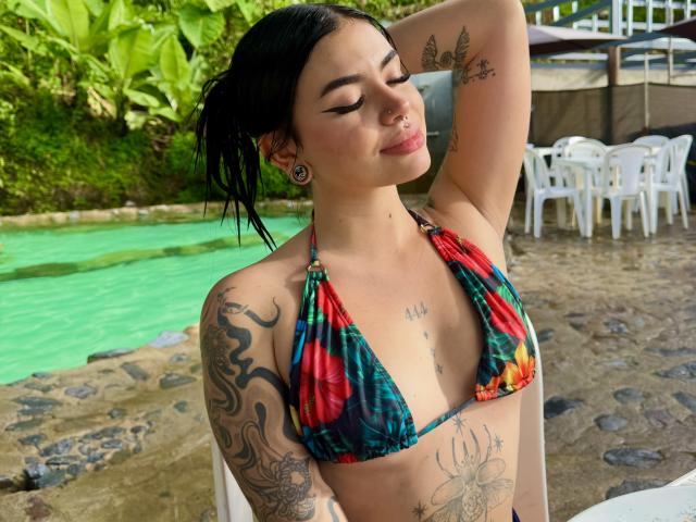 VioletteVanz with tattoos, dark hair in a ponytail, wearing a colorful bikini, eyes closed, arm raised, showing off her tits and piercings, sitting by a green-tinted pool.