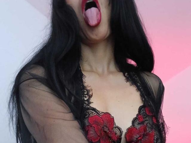 ZarayFeet with long black hair, tongue out, wearing a sheer black lace top with red floral embroidery, exposing her cleavage and red lipstick.