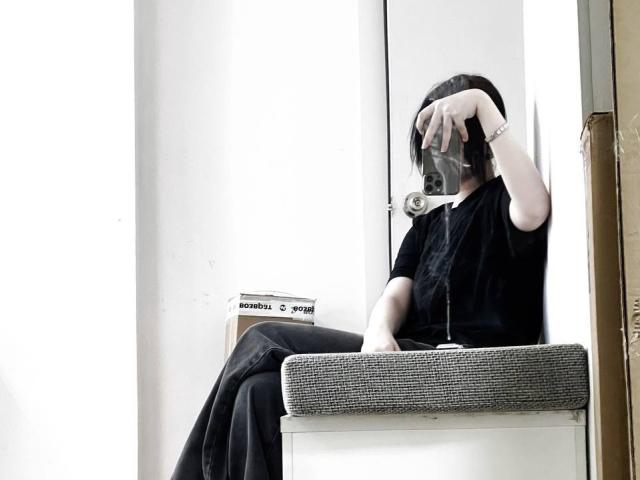 MariannaWarren, with black hair, black shirt, and black pants, sits on a grey upholstered chair, holding a phone, covering her face, with a wristwatch on her left wrist.