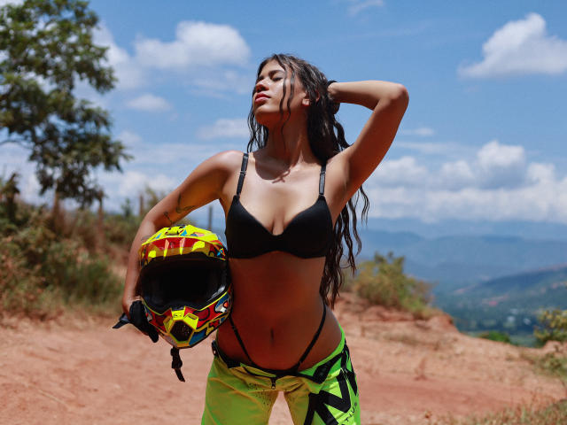 NikaFoxxyHot with long black hair, black bra, neon green pants, holding a yellow and red helmet, standing on red dirt, hand in hair, looking up, clear blue sky.