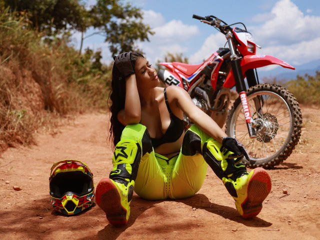 NikaFoxxyHot sits on dirt, legs spread, neon pants, black top, red boots, motorcycle behind, helmet on ground, smiling, black bra, curvy, sexy, tan skin, brown hair.