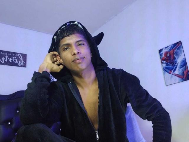 MickStormx, ein junger Mann mit kurzen, schwarzen Haaren, der einen schwarzen Kapuzenpullover mit Katzenohren trägt, sitzt auf einem Bett, lächelt leicht und schaut in die Kamera, in einem blau beleuchteten Raum..