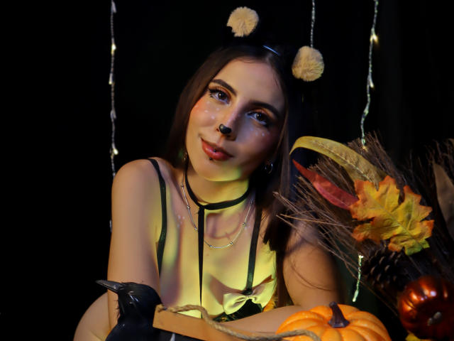 NovaLewis, con cabello castaño, orejas de gato, nariz negra, ojos brillantes, vistiendo un collar negro, una blusa amarilla, sosteniendo una calabaza y un guante negro, rodeada de decoraciones otoñales..