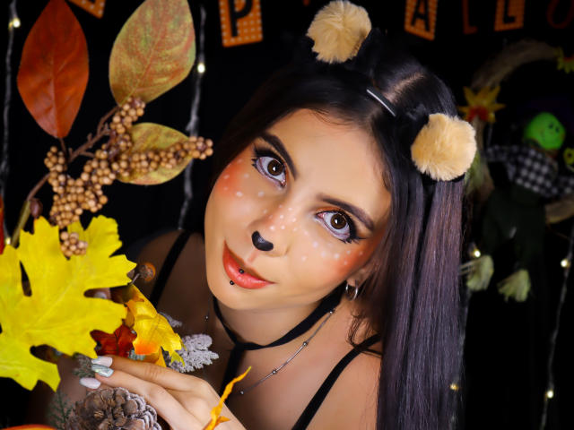 NovaLewis, com cabelo escuro, orelhas de urso, nariz preto e sardas, segurando folhas amarelas, tem o lábio perfurado, usa um colar preto e tem grandes olhos castanhos, em uma foto fofa com tema de outono..