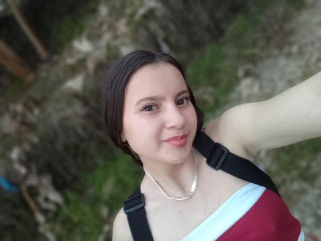 BeccaSimons with short brown hair, light skin, small tits, wearing a red and white top, black straps, and a silver necklace, smiling in a selfie.