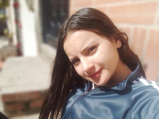 BeccaSimons with wet black hair, smiling at the camera, wearing a blue Adidas jacket, her face and small nose highlighted in the shot. Her tits are not visible.