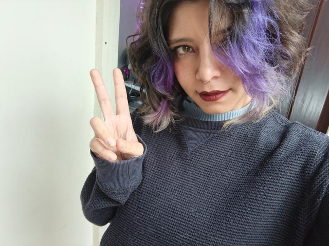 DarckVelvet with purple and grey curly hair, wearing a dark navy knitted sweater, blue choker, and dark red lipstick, making a "peace" sign with their right hand.