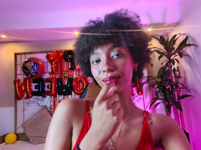 KiaraKamau with curly black hair, red lingerie, finger in mouth, pink and purple lighting, and a sexy smile. Boobies visible in the red top. Potted plant in the background.