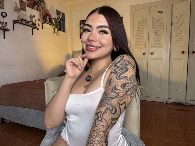 VioletteVanz with long dark hair, smiling, wearing a white tank top, showing tattooed arm, touching chin, sitting on chair, brown bed in background, white closet doors.