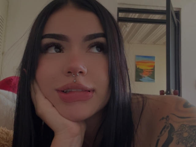 VioletteVanz with long black hair, nose ring, lip piercing, tattoo on right arm, resting cheek on hand, looking up with dark eyebrows and pink lipstick.