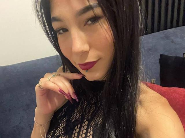 VioletaVelvet with long black hair, dark red lipstick, and black fishnet top, showing off manicured nails and rings, winking and smiling at the camera.