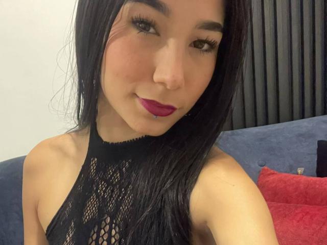 VioletaVelvet has long black hair, light skin, purple lipstick, and a black mesh top, showing her small tits and a slight smile with a lip piercing.