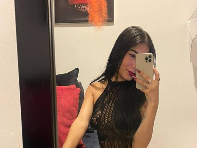 VioletaVelvet taking a mirror selfie, wearing a black mesh halter top, long black hair, red lipstick, and holding a gold iPhone. Her tits are visible through the mesh.