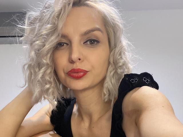 DouceAmelia with curly blonde hair, red lipstick, black lace top, and a slight smirk, looking directly at the camera. Her tits are small and her ass is not visible.