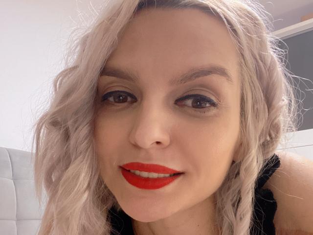 DouceAmelia has light blonde, wavy hair, red lipstick, and is smiling with dark, smoky eyes. She's wearing a black top, and her face is centered in the photo.