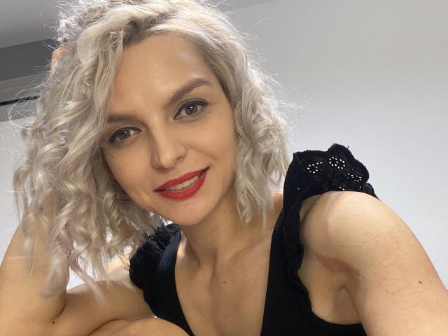 DouceAmelia with curly white hair, red lipstick, black dress, smiling at the camera, showing her tits, and holding her head with one hand.