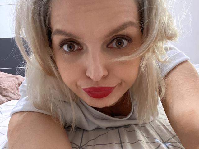 DouceAmelia with blonde hair, red lipstick, and brown eyes, looking at the camera with a slight smile, wearing a white shirt, lying on a bed with a striped sheet.
