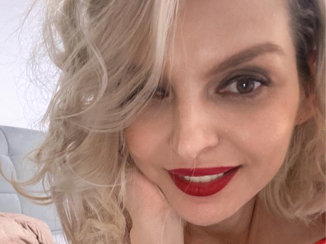 DouceAmelia with blonde curly hair, red lipstick, and brown eyes, smiling softly, hand resting near her face, showing off her sexy face and cute nose.