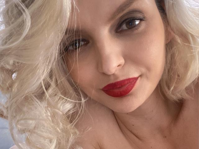 DouceAmelia has blonde curly hair, red lipstick, and a slight smile, with brown eyes looking at the camera. Her bare shoulders are visible.